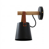 A modern wall-mounted sconce with a matte black cone-shaped shade, a natural wood horizontal arm, a black round wall plate, and a brown leather strap detail.
