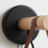 Wall sconce close-up shows a round black wall plate, natural wood peg and brown strap against a white wall.