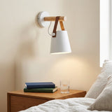 White wall sconce with a wooden peg and brown straps shines above a bedside table holding books and a glass.