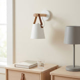 A wall-mounted sconce with a white conical shade, light wood rod, and brown leather straps, shown lit in a softly styled room beside a gray table lamp and a light wood bedside table.