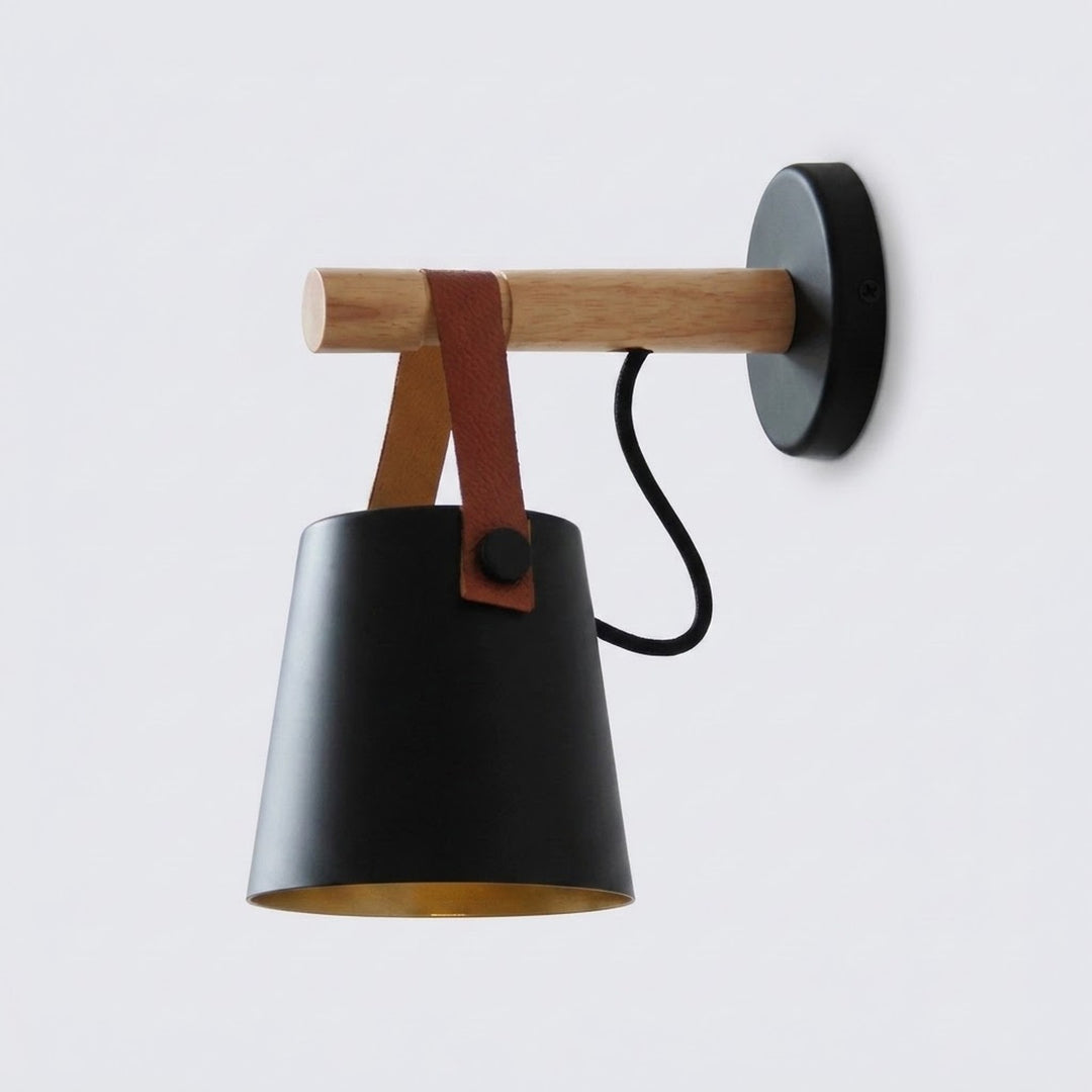 A modern wall-mounted sconce with a matte black metal cone shade, a natural wood horizontal arm, a brown leather strap detail, and a black circular wall plate, shown against a plain light gray background.