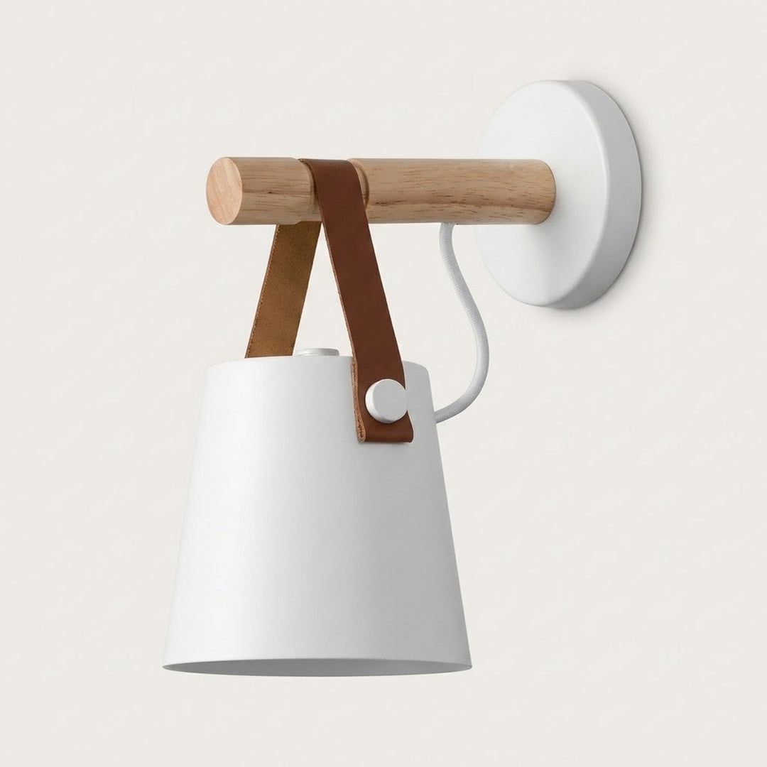 White wall sconce with a tapered shade, natural wood peg, brown straps and round white wall plate against a pale background.