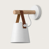 White wall sconce with a tapered shade, natural wood peg, brown straps and round white wall plate against a pale background.