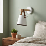 Unlit white wall sconce with wood and brown straps hangs on a sage wall above a nightstand with a plant and cup.
