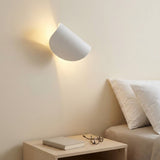 A white wall sconce casts warm light across a beige bedroom wall above a bedside table beside white pillows.