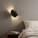 A modern black wall-mounted sconce lamp glowing with warm light on a gray bedroom wall above a wooden bedside table and bed.