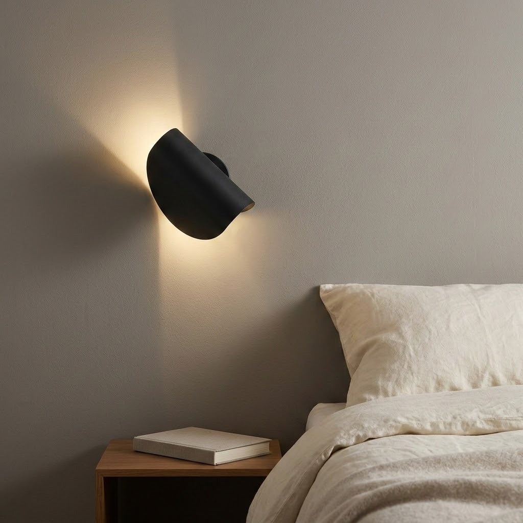 A modern black wall-mounted sconce lamp glowing with warm light on a gray bedroom wall above a wooden bedside table and bed.