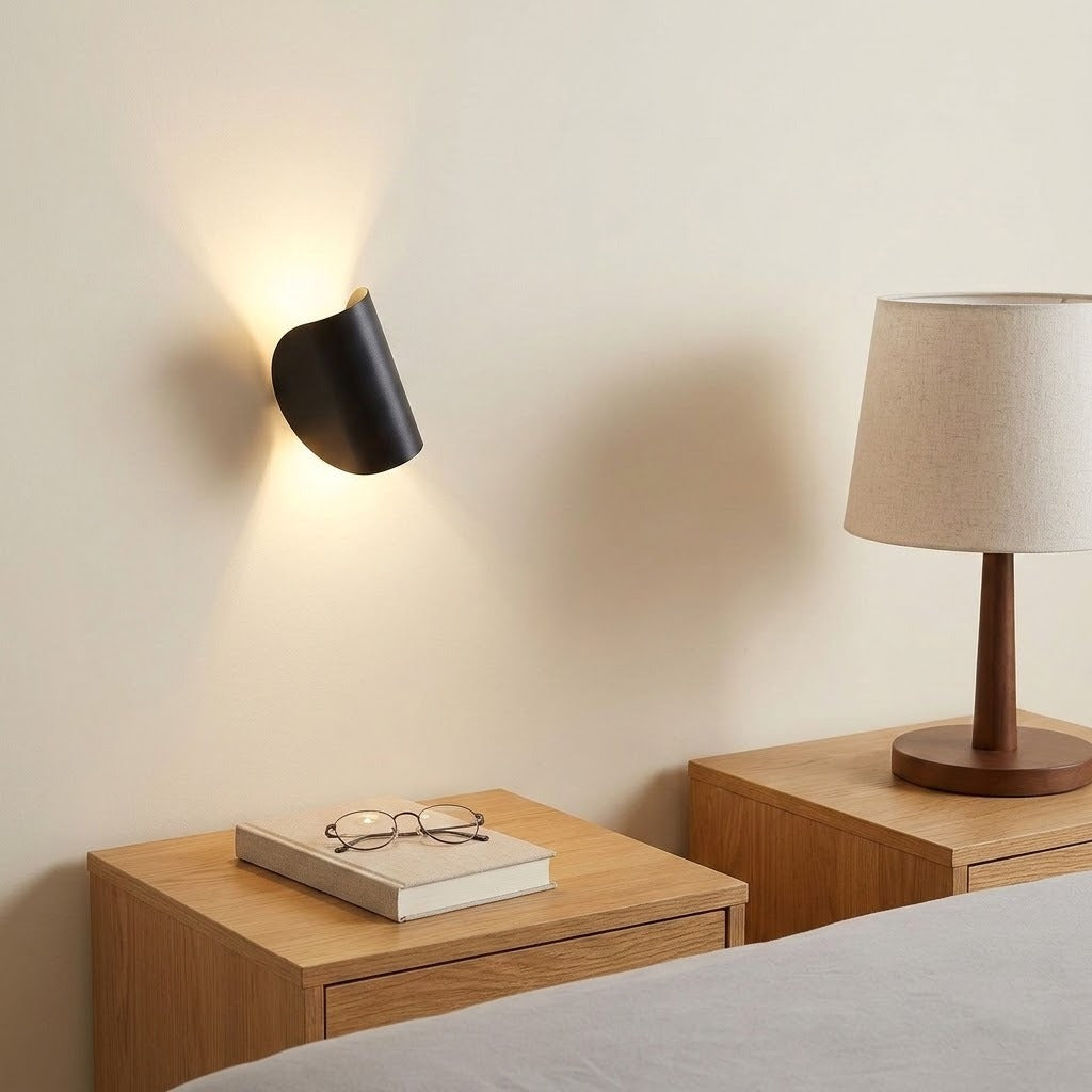 A modern black wall-mounted sconce lamp glowing warmly on a beige wall above a wooden bedside table, with a lamp and book visible in the room setting.