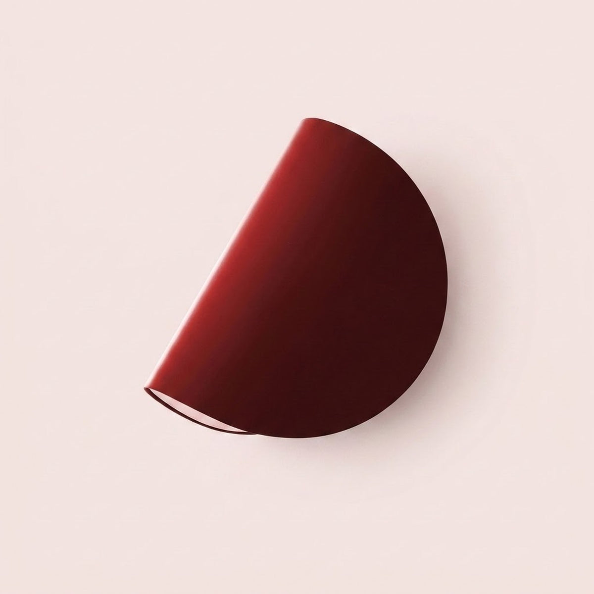 A modern semi-circular wall sconce in a deep burgundy red finish, mounted on a solid pale pink wall.