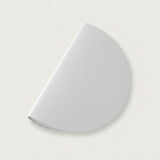 A white wall sconce appears diagonally against an off-white background, showing its semicircular shade and a subtle shadow.