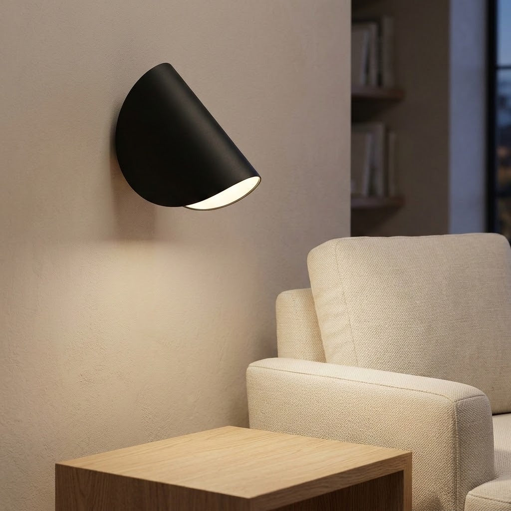A modern black semi-circular wall sconce light fixture mounted on a beige wall, casting a warm glow downwards next to a cream-colored armchair and a light wood side table.