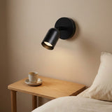 A black wall sconce with an adjustable cylindrical shade glows warmly above a wooden bedside table in a cozy bedroom.
