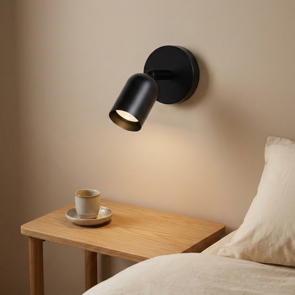 A black wall sconce with an adjustable cylindrical shade glows warmly above a wooden bedside table in a cozy bedroom.