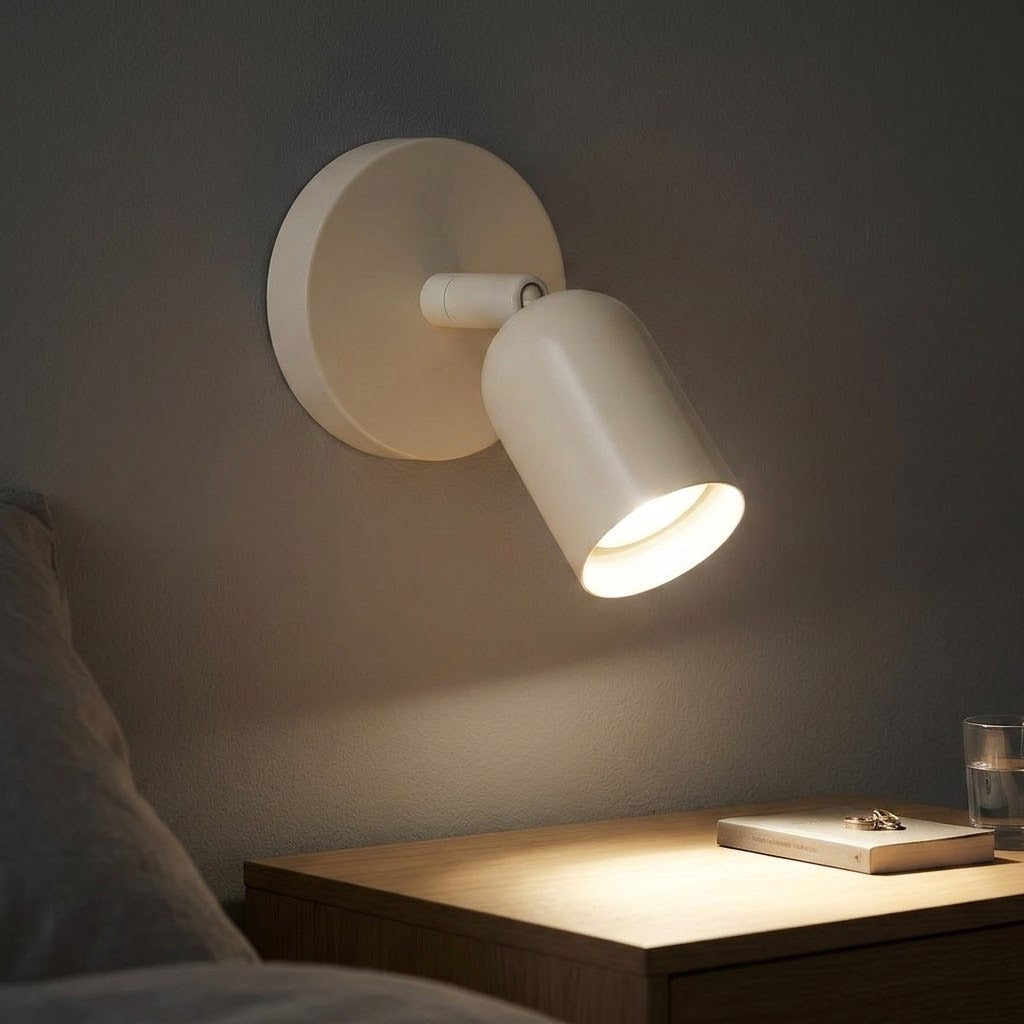 A cream wall sconce with an adjustable shade casts warm light over a wooden bedside dresser in a dark bedroom.