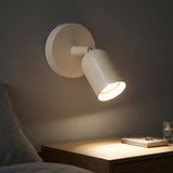 A cream wall sconce with an adjustable shade casts warm light over a wooden bedside dresser in a dark bedroom.