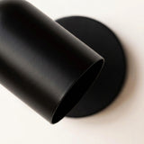 Close-up of the matte black metal shade and round wall plate of a wall sconce on an off-white background.