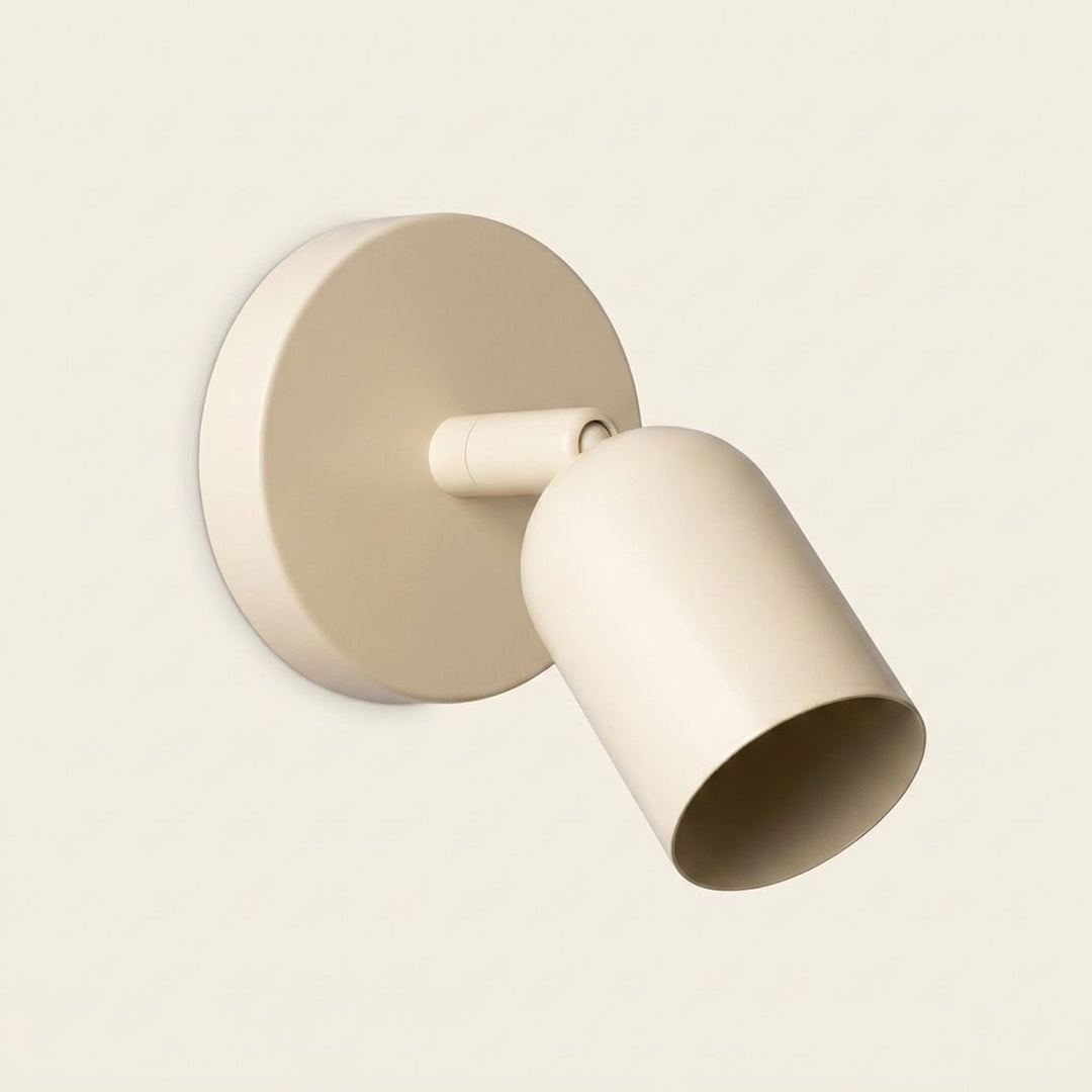 A cream wall sconce with a round backplate and downward angled shade has a dark opening against a pale cream background.