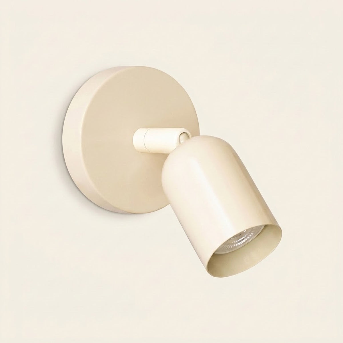 A cream wall sconce with a round backplate and downward angled shade has a visible bulb against a pale cream background.