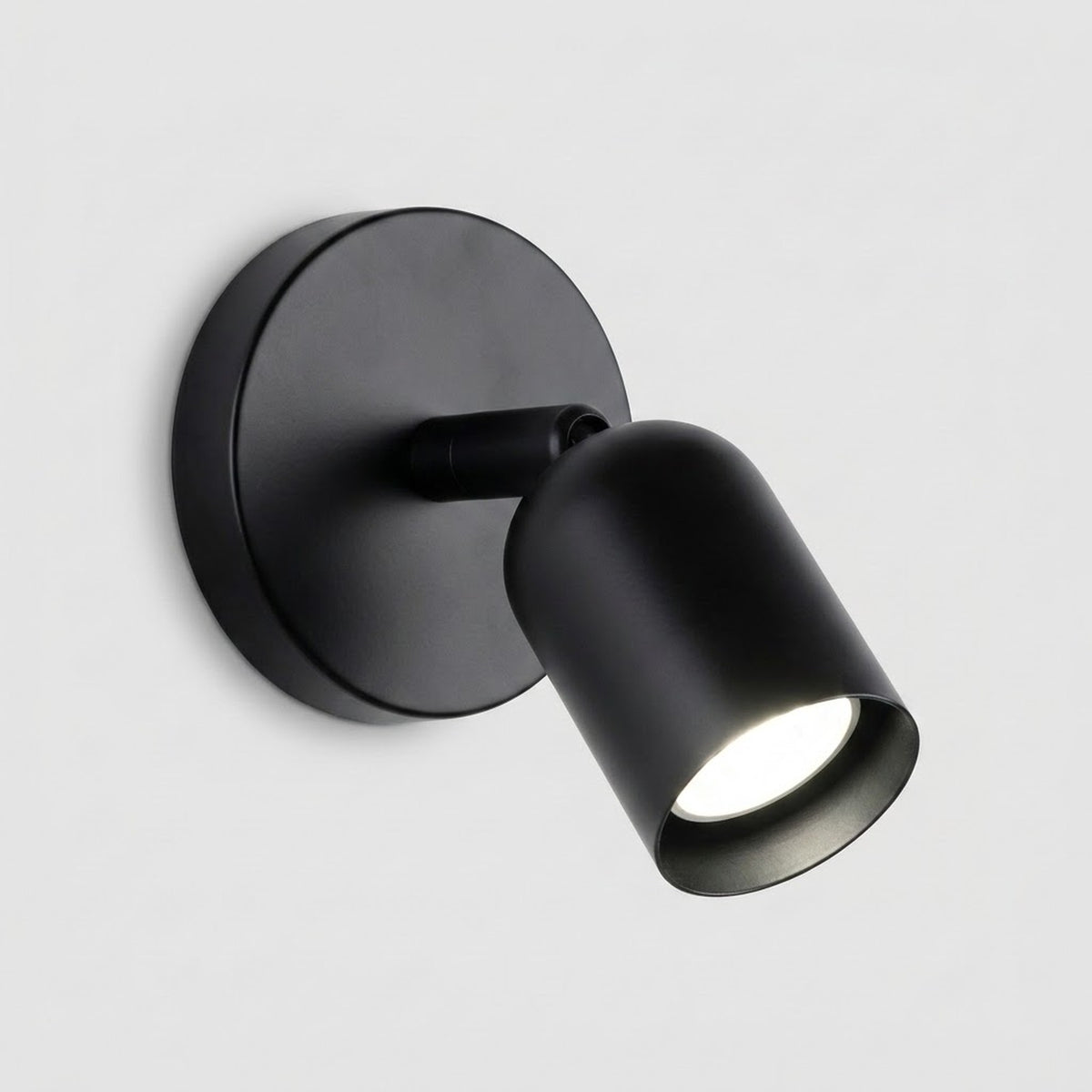A matte black wall sconce with a round backplate and downward angled shade emits white light against a light grey background.