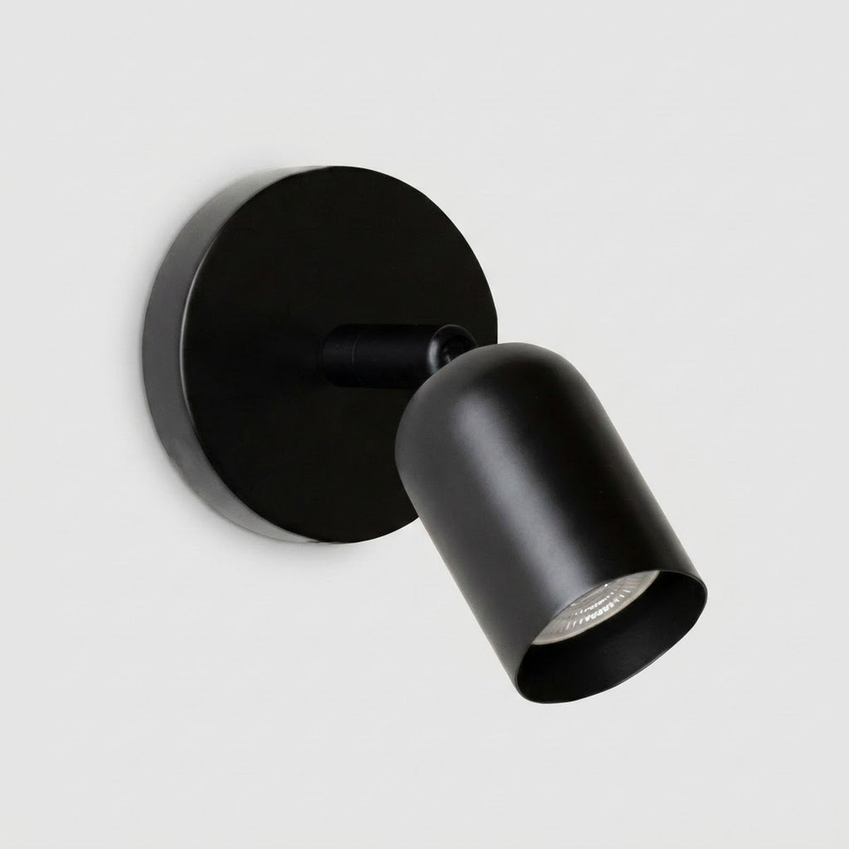 A modern matte black spotlight wall sconce with a circular backplate and adjustable cylindrical shade, shown against a light gray background.