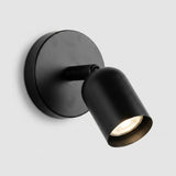 A matte black wall sconce with a round backplate and downward angled shade glows warmly against a light grey background.