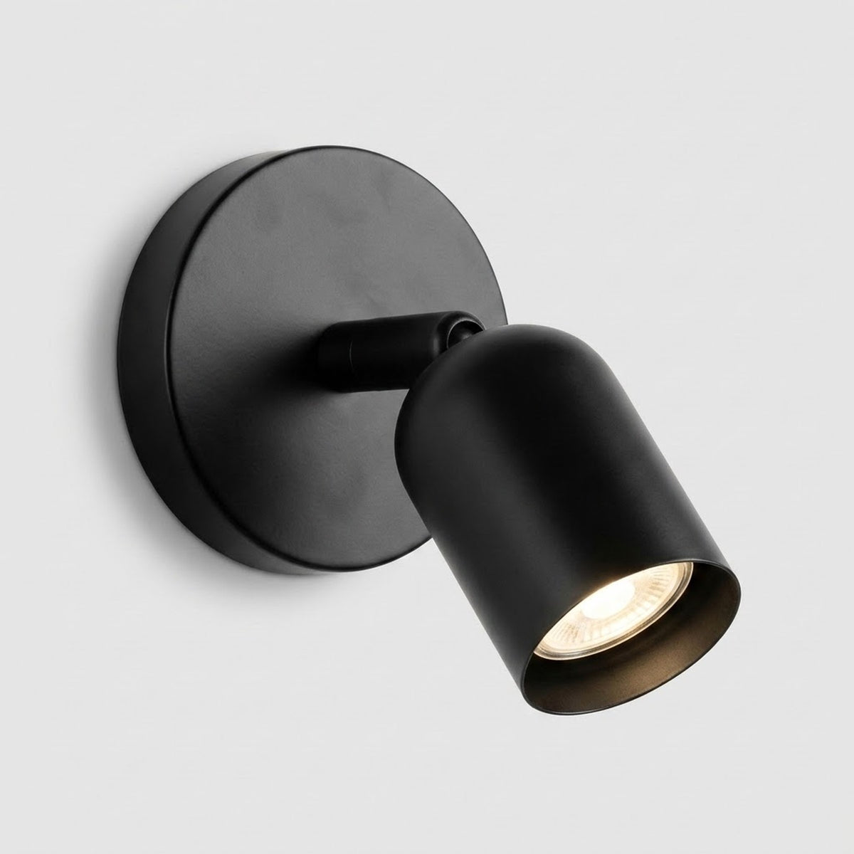 A matte black wall sconce with a round backplate and downward angled shade glows warmly against a light grey background.