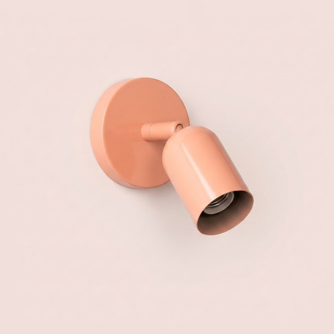 Peach-pink wall sconce with an empty bulb socket inside its downward-angled shade against a pale pink background.