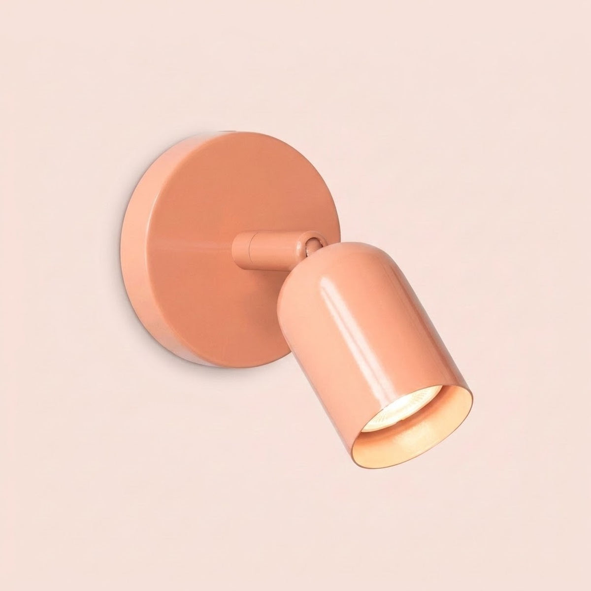 Peach-pink wall sconce with a round backplate and glowing bulb against a pale pink background.