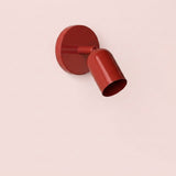 Glossy red wall sconce with a cylindrical shade angled downward and a round backplate against a pale pink background.