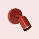 Glossy red wall sconce with a circular backplate and downward-angled shade against a pale pink background.