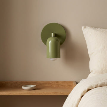 An olive green wall-mounted sconce with a circular backplate and a glossy cylindrical shade, shown installed above a wooden bedside table next to a bed with white bedding.