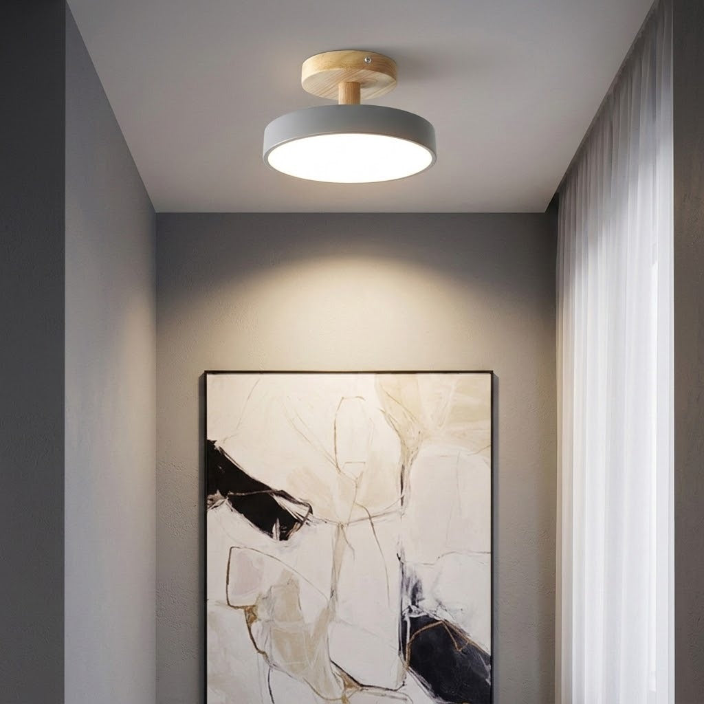 A modern semi-flush mount ceiling light with a circular white shade and a light wood ceiling canopy, mounted on a white ceiling in a hallway above a framed abstract painting.