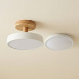 A modern ceiling light fixture shown from below, featuring a circular white shade with a natural wood ceiling mount and a second matching round light element to the right, set against a plain beige wall.