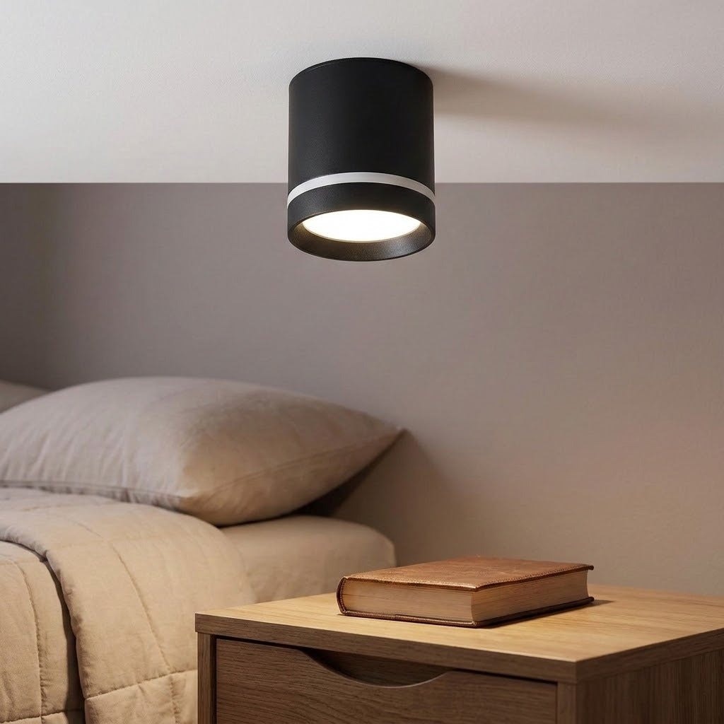 Black cylindrical LED downlight with a silver-colored band glows above a bedside table beside a bed.
