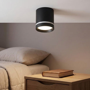 Black cylindrical LED downlight with a silver-colored band glows above a bedside table beside a bed.