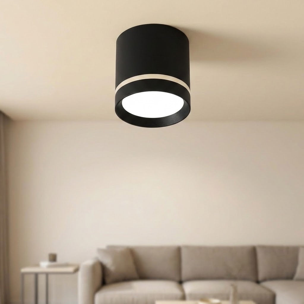 Illuminated black cylindrical LED downlight with a silver-colored band is mounted on a pale ceiling above a beige sofa.