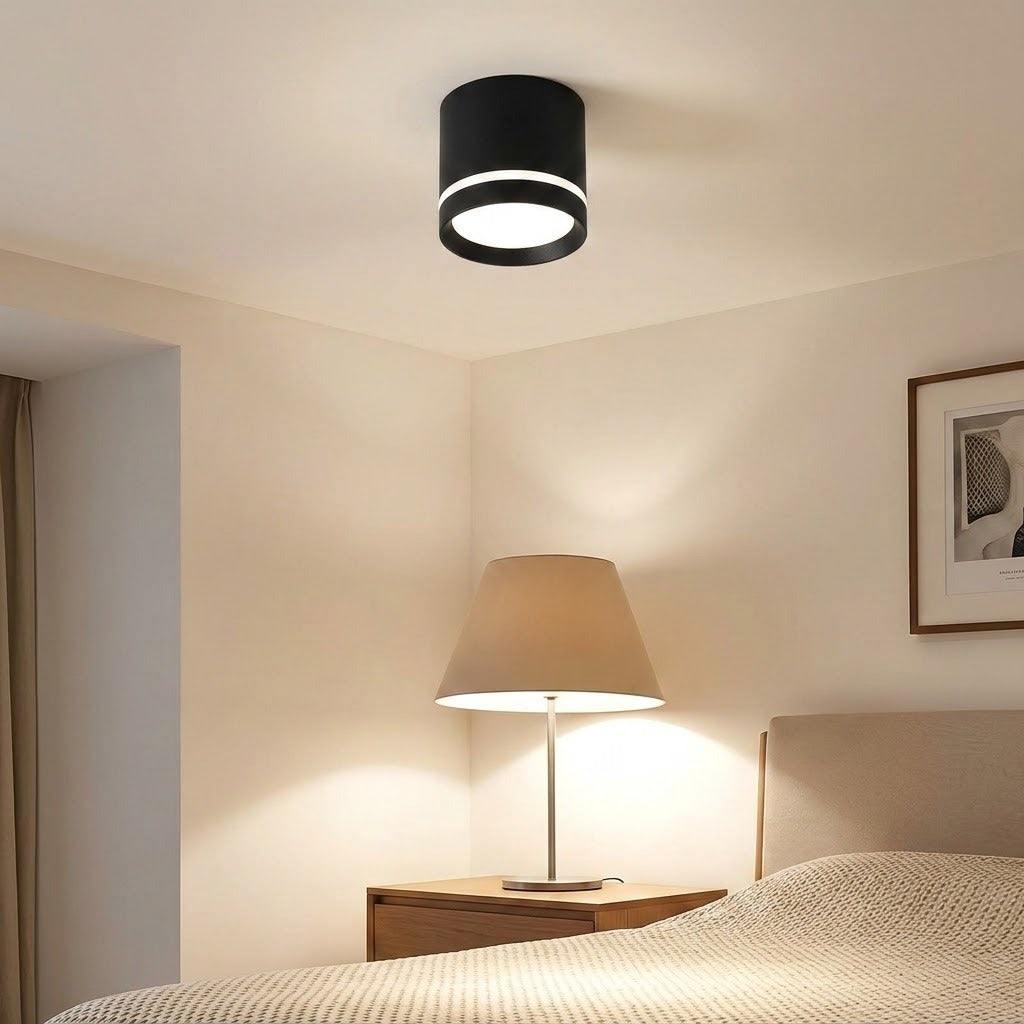 A black cylindrical LED downlight with a bright accent ring illuminates a beige bedroom with a bedside lamp.