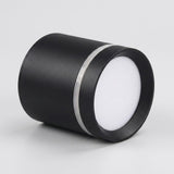 A black cylindrical LED downlight rests on a light gray surface, with its white diffuser facing right and silver trim visible.