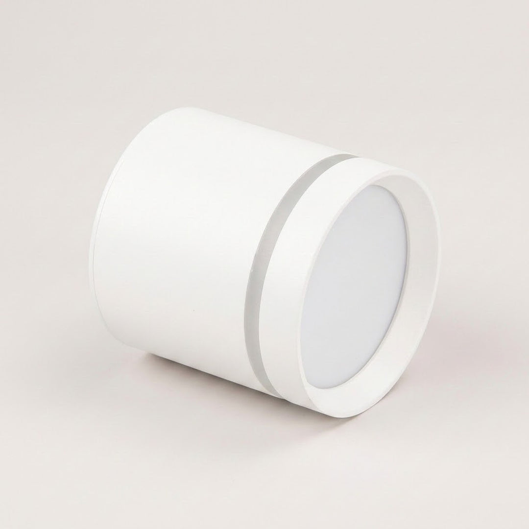 A white cylindrical LED downlight rests on a pale beige background, showing its recessed circular diffuser and silver-colored accent ring.