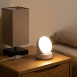 A small round white night light with a glowing illuminated face sits on a wooden bedside table next to a larger table lamp with a beige fabric shade and black base in a bedroom setting.
