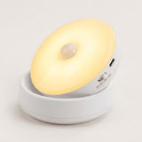 A small white portable LED night light with a circular glowing lamp and a built-in motion sensor, shown on a clean light gray background.