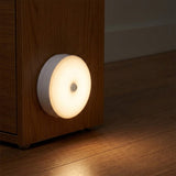 A round white smart night light glowing with warm white light, shown mounted on a wooden cabinet next to a wood floor.