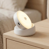 A small round white LED night light with a glowing face, resting on a light wood bedside table in a softly lit bedroom setting.