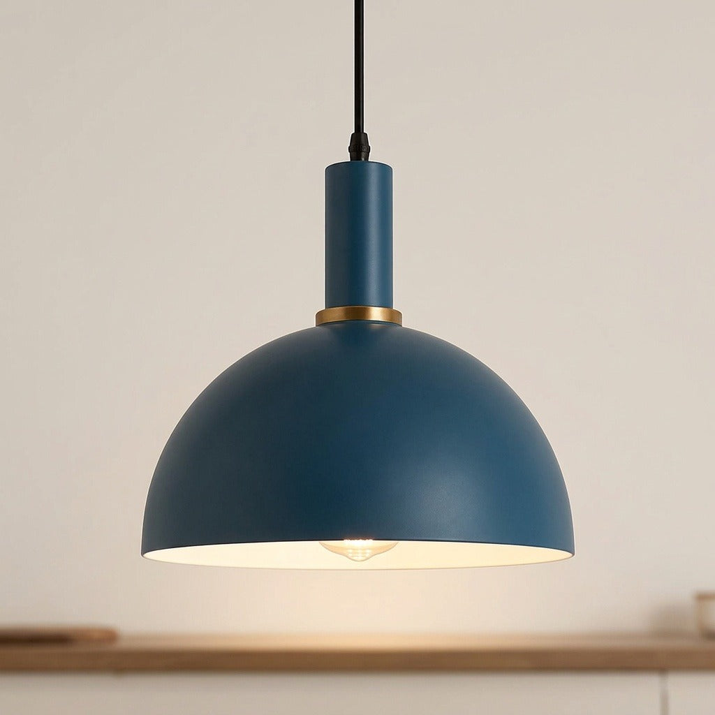 A modern dome-shaped pendant light in matte blue with a gold accent ring and a black hanging cord, shown illuminated against a warm off-white background.