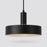 A modern black pendant light with a wide cylindrical shade, a matte black metal body, and a warm cream-colored interior, suspended against a light gray background.