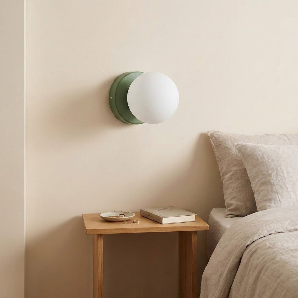 A wall sconce with a green iron base and white plastic globe shade sits above a wooden bedside table beside neutral bedding.