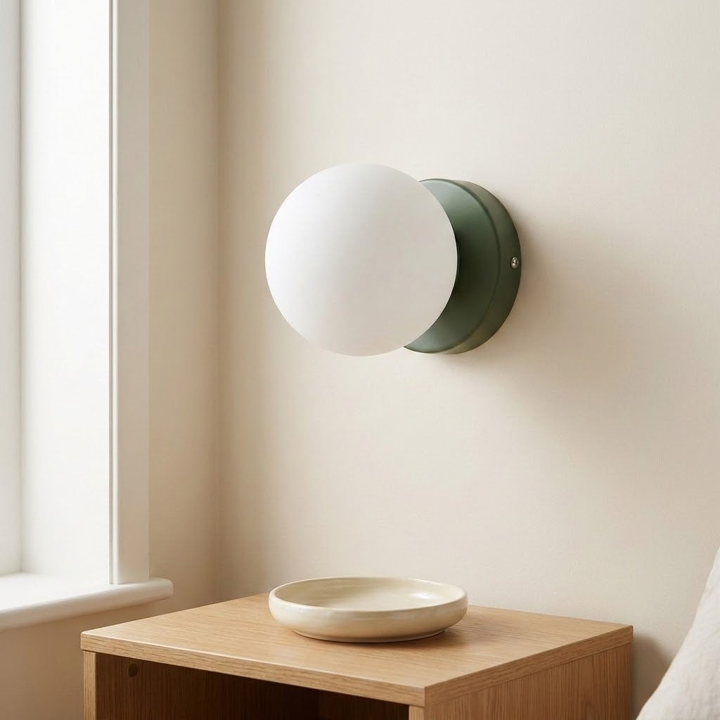 A wall sconce with a green iron base and white plastic globe shade is mounted beside a window above a wooden cabinet.