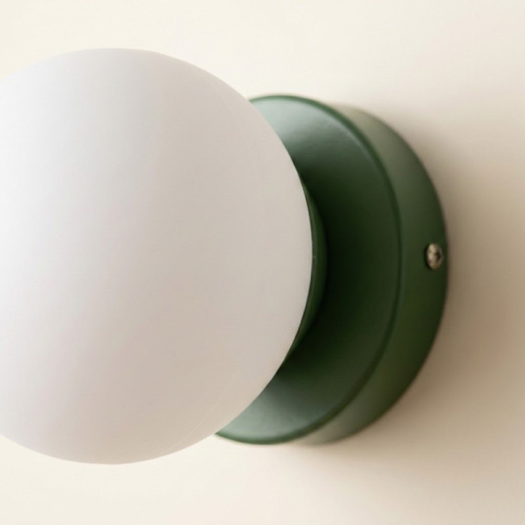 A close-up shows the wall sconce’s green iron base, visible side screw, and white plastic globe shade against a cream wall.