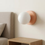 A wall sconce with a peach-pink iron base and white plastic globe shade sits above a wooden bedside table beside an upholstered headboard.