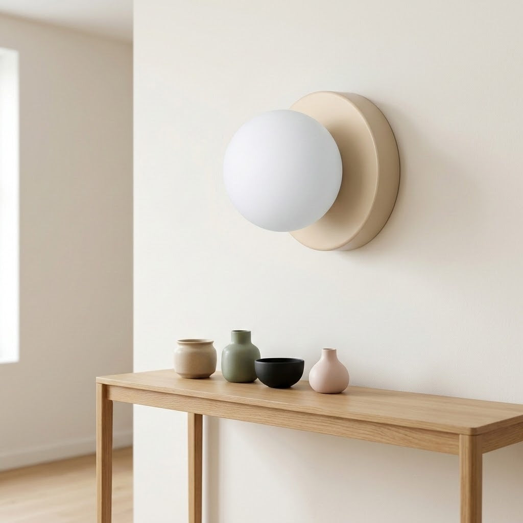 Wall sconce with a white globe shade and beige circular base mounted above a wooden console displaying small vases and a bowl.
